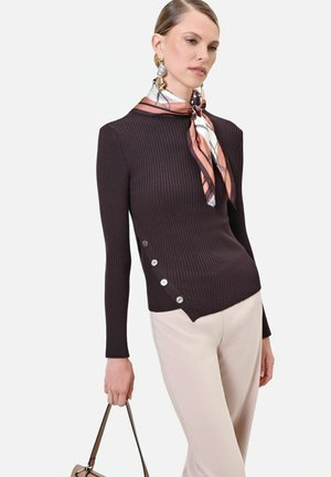 Donna che indossa un maglione a coste scuro con bottoni laterali, pantaloni chiari, una sciarpa di seta con fantasia intorno al collo, grandi orecchini e tiene una borsa per la maniglia.