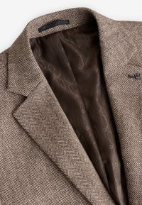 Brauner Blazer mit Fischgrätmuster und einem glatten braunen Innenfutter. Verfügt über geschlitzte Revers und ein dezentes Logodetail auf der Vorderseite.