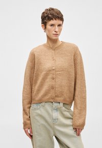 Καφετί πλεκτό κάρντιγκαν με στρογγυλή λαιμόκοψη, μακριά μανίκια και έξι κουμπιά μπροστά, φορεμένο πάνω από ανοιχτό μπλε τζιν παντελόνι.