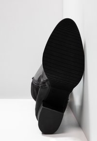 Bottine noire à chevilles avec une surface en cuir lisse, une semelle épaisse texturée et un talon modeste, inclinée sur un fond neutre.