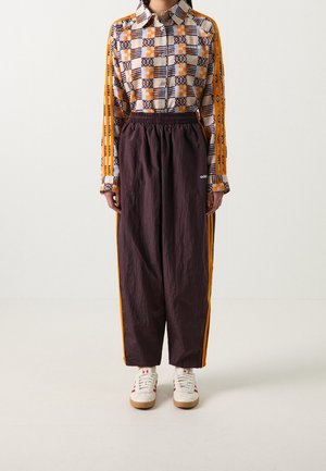 Osoba nosząca wzorzystą koszulę na guziki z długim rękawem z pomarańczowymi paskami oraz ciemnofioletowe spodnie dresowe Adidas z pomarańczowymi paskami po bokach i białe tenisówki.