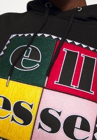 Černá mikina s kapucí se skládá z patchworkového designu s zelenými, červenými, žlutými a růžovými čtverci, z nichž každý má tučný, kontrastní text v černé barvě.