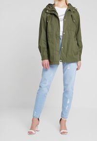 Olive green lightweight jacket with a hood, side pockets, button details, and a drawstring waist, worn over a white graphic t-shirt and blue jeans.