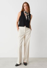 Zwart cropped vest met knoopsluiting, gecombineerd met hoge crème broek. Model draagt een zwart-wit gestreepte sjaal en platte schoenen.