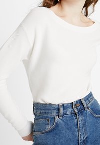 Vit ribbad långärmad tröja med vid halsringning, bärs med högmidjade blå denimjeans med knappgylf och klassisk femficksdesign.