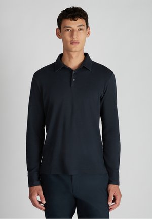 Man in een zwart langslavend polo-shirt staat tegen een effen lichtgrijze achtergrond.