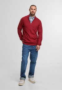 Czerwony sweter z zamkiem na szyi, noszony na niebieskiej koszuli w kratę, w zestawieniu z podwiniętymi niebieskimi dżinsami i białymi trampkami. Miękka tekstura, casualowy design.