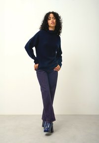 Dunkelblauem Strickpullover mit hohem Kragen, kombiniert mit lila ausgestellten Jeans und glänzenden blauen Absatzstiefeln. Glatte Stofftexturen und figurbetonte Formen.