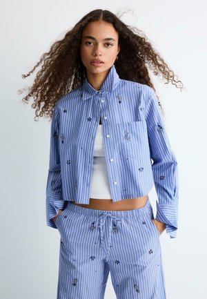 Mlada žena s kovrčavom kosom koja nosi plavo-bijelu prugastu izvezenjak jaknu i odgovarajuće hlače, ruke u džepovima, na jednobojnoj pozadini.