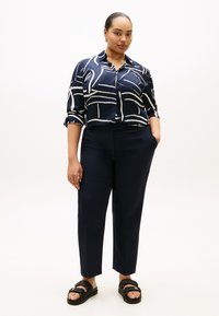 Μπλούζα σκούρου μπλε ναυτικού χρώματος με λευκό αφηρημένο σχέδιο, κουμπωτή στο μπροστινό μέρος και με μανίκια που έχουν αναδιπλωθεί· συνδυασμένη με προσαρμοσμένο παντελόνι ναυτικού χρώματος και μαύρα σανδάλια.