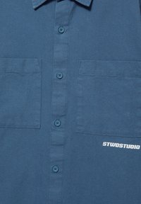 Chemise bleue boutonnée avec deux poches poitrine et texte blanc "STWDSTUDIO" sur la poche inférieure droite.