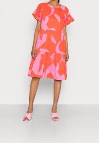 Vestido rojo brillante y rosa con mangas con volantes, diseño en capas y grandes patrones geométricos. Combinado con zapatos abiertos rosa.