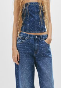 Top corto in denim e jeans a gamba larga, entrambi di un blu scuro con macchie bianche sparse. I jeans hanno tasche frontali e chiusura con bottone.