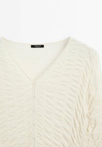 Cardigan crème texturé avec un col en V, présentant un froncé vertical, une fermeture avant à boutons, et un tissu en maille lisse. Étiquette de marque visible.