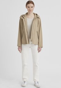 Beige wasserdichte Jacke mit Kapuze, Reißverschluss vorne und zwei Seitentaschen, getragen über einem weißen Oberteil und kombiniert mit weißen Hosen und Sneakers.