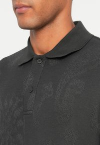 Mann mit unterem Gesicht und Hals, der ein dunkelgraues, geknöpftes Polohemd mit einem dezenten floralen Strickmuster trägt.