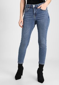 Džíny s vysokým pasem a slim fit v středně modrém denimu, se třemi kapsami, decentním šitím a ohrnutými lemy, doplněné černými kotníkovými botami.