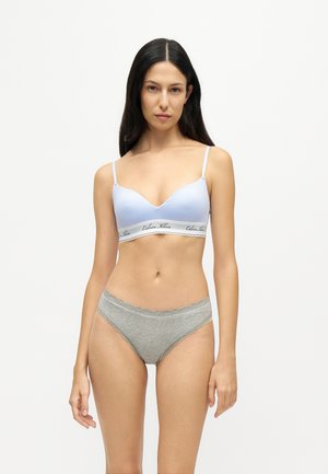 Femme aux longs cheveux foncés portant un soutien-gorge bleu clair avec bande logo Calvin Klein et une culotte grise bordée de dentelle, debout sur fond blanc.