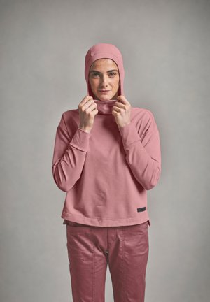 Frau trägt ein pinkfarbenes, langärmliges Kapuzenshirt und eine pinke Hose, richtet mit beiden Händen die Kapuze vor einem einfarbigen grauen Hintergrund.