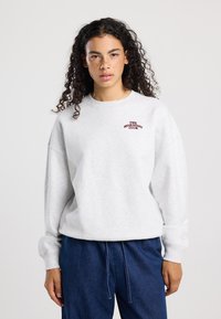 Lichtgrijze sweatshirt met ribgebreide ronde hals, oversized pasvorm en geborduurd "THE MORNING CLUB" in donkerrood. Gecombineerd met donkerblauwe broek.