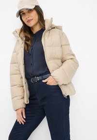 Vrouw in een beige pufferjack, marineblauw knopenhemd, donkere jeans met een zwarte riem en een beige pet, staande tegen een witte achtergrond.
