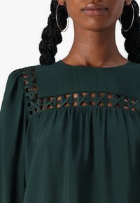 Blusa verde con tessuto strutturato, dotata di un design decorativo a taglio sul giogo e maniche morbide e arricciate.