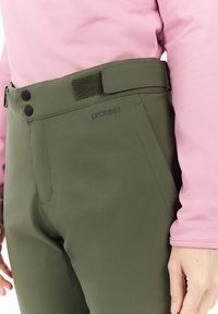 Olijfgroene broek met een strak design, voorzien van een platte tailleband met twee zwarte knopen en een klittebandaccent, gecombineerd met een roze langemouwtop.