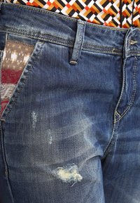 Denimjeans i mörkblått med en sliten textur. Har synligt slitage med lappar av flerfärgad tyg på en ficka.