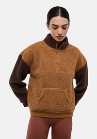 Pull en polaire marron avec un col à demi-fermeture éclair, des manches foncées contrastées, une poche avant et une surface texturée.