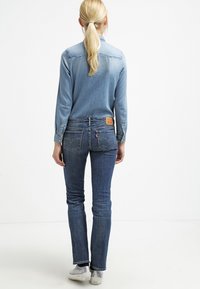 Camisa de mezclilla azul clara, de manga larga, y jeans azul oscuro con una etiqueta de cuero color marrón claro. Silueta ajustada, costuras sutiles y bolsillos traseros visibles.