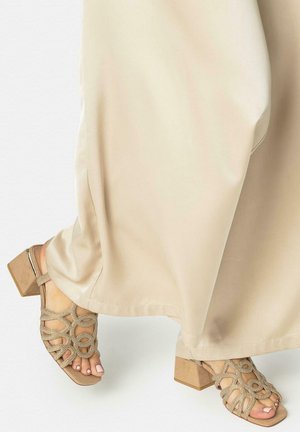 Pies con sandalias beige de tacón ancho y punta abierta, con tiras decorativas, combinadas con pantalones largos de color crema.