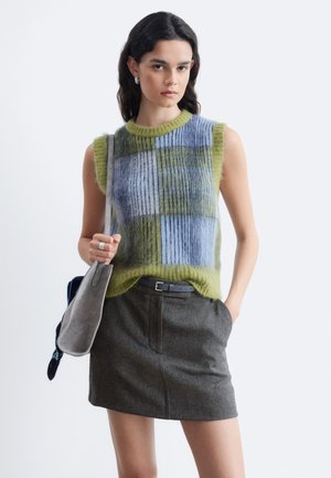 Frau trägt einen ärmellosen, flauschigen karierten Pullover in Blau und Grün mit einem grauen Rock, hält eine graue Schultertasche und steht vor weißem Hintergrund.