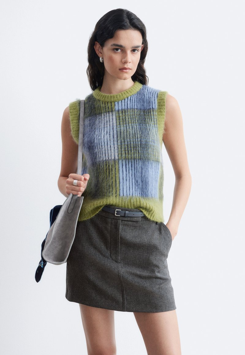 Femme portant un pull sans manches à carreaux duveteux bleu et vert avec une jupe grise, tenant un sac à épaule gris, debout sur un fond blanc.