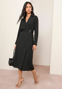 Schwarzes Wickelkleid mit langen Ärmeln, kragenähnlichem Ausschnitt, silberner Schnalle in der Taille und knielangem Saum. Das Model trägt kettenartige Absätze.