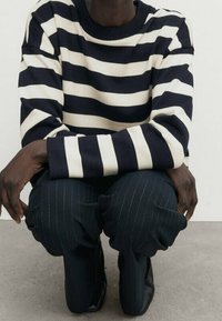 Personne accroupie portant un pull à rayures horizontales noires et blanches et un pantalon à fines rayures foncées avec des chaussures noires sur un sol gris.