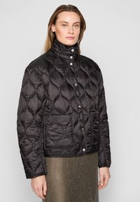 Black quilted jacket with a high collar, featuring large diamond patterns, snap buttons, and side pockets. Paired with a metallic skirt.
