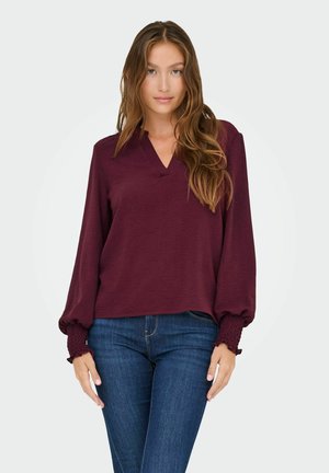 Ung kvinde med langt bølget hår iført en bordeaux langærmet bluse med elastiske manchetter og blå jeans, der står mod en ensfarvet baggrund.