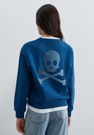 Persona con cabello largo y oscuro lleva una sudadera azul con un gran diseño de calavera y huesos cruzados en la espalda sobre vaqueros de mezclilla claros.
