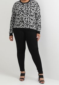 Osoba ubrana w czarno-białą bluzkę w panterkę z długim rękawem, czarne dopasowane spodnie oraz czarne sandały na słupku z odsłoniętymi palcami.