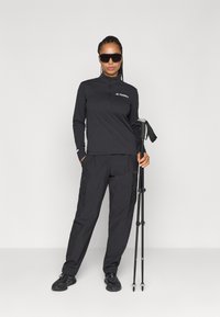Svart sportig långärmad topp med dragkedja vid kragen och logotyp, i kombination med matchande cargo-byxor. Håller ett vandringsstavar. Solglasögon fulländar utseendet.