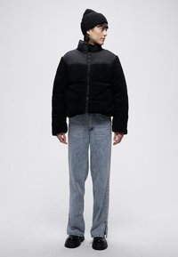 Černá mikina z fleecu a izolovaná bunda se zapínáním na zip vpředu, doplněná světlými modrými džínami a černými robustními botami; model nosí černou čepici.