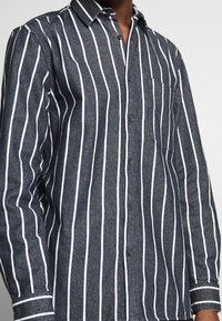 Camisa de botones gris oscuro con rayas verticales blancas, que presenta un bolsillo en el pecho y un colla clásico. Tela texturizada, de manga larga.