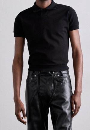 Polo noir à manches courtes, coupe slim, associé à un pantalon en cuir noir taille haute porté par une personne debout sur un fond uni.