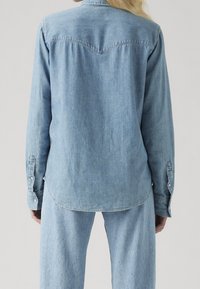 Vaaleansininen denimpaita, jossa pitkä hihan, painonapillinen, kaareva helma ja länsityylinen yökki selässä. Tasainen pinta kauttaaltaan.