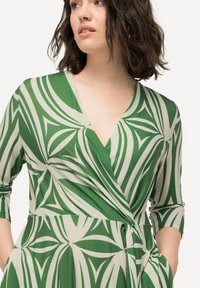 Grünes Wickelkleid mit einem auffälligen Blattmuster in Weiß, 3/4-Ärmeln und V-Ausschnitt. Aus leichtem, glattem Stoff mit Seitentaschen gefertigt.
