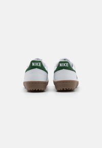Białe sneakersy z zielonymi akcentami i wszytymi wstawkami na piętach, z teksturowaną brązową gumową podeszwą oraz wyraźnym logo Nike na piętach.