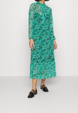 Femme portant une robe midi verte à fleurs, manches longues, col lavallière et mocassins noirs avec un détail chaîne dorée.