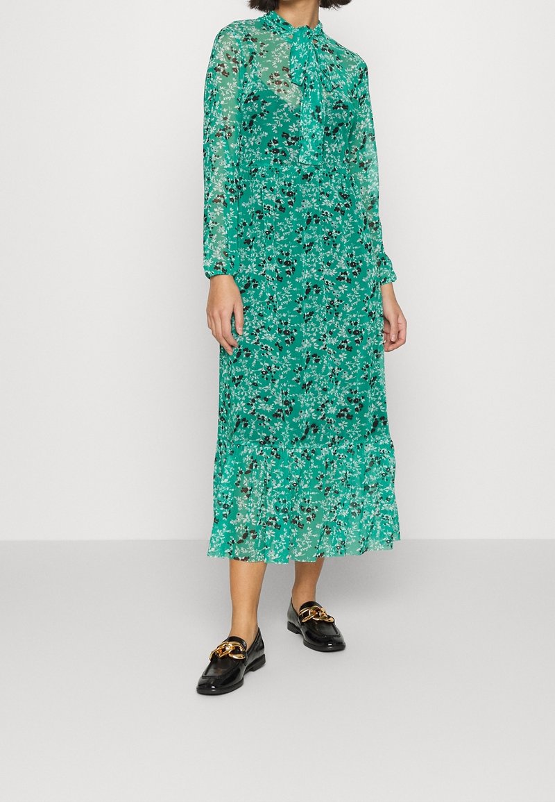 Femme portant une robe midi verte à fleurs, manches longues, col lavallière et mocassins noirs avec un détail chaîne dorée.