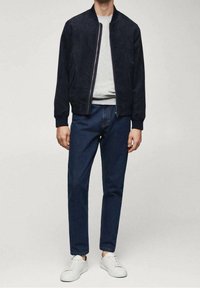 Giacca bomber in camoscio blu navy, maglietta grigia con scollo rotondo, pantaloni in denim scuro e sneaker bianche, con chiusura a zip e polsini a costine.