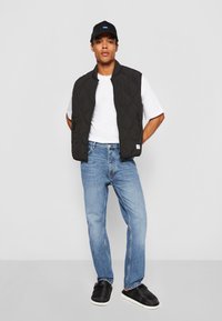 Black quilted vest over a white t-shirt, paired with light blue jeans and black slip-on shoes. The cap is black with a blue logo.
